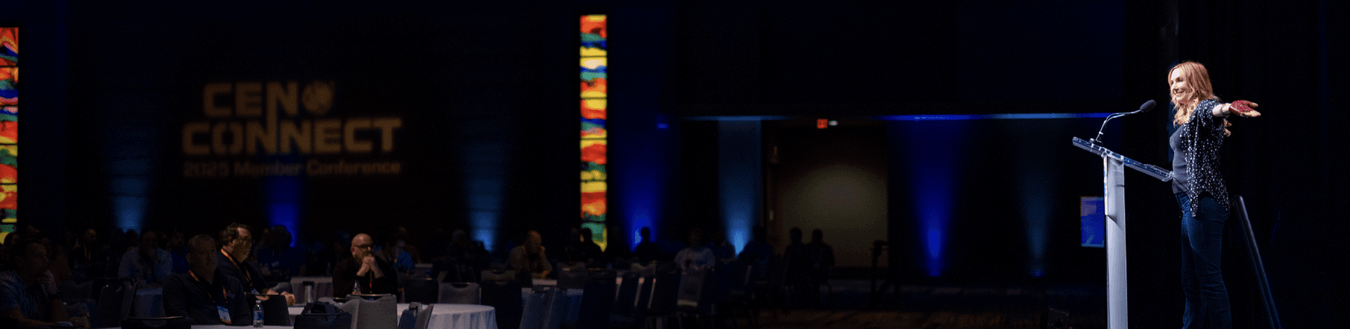 Keynote speaker on stage with light shown on them and a crowd at attendees in the audience.